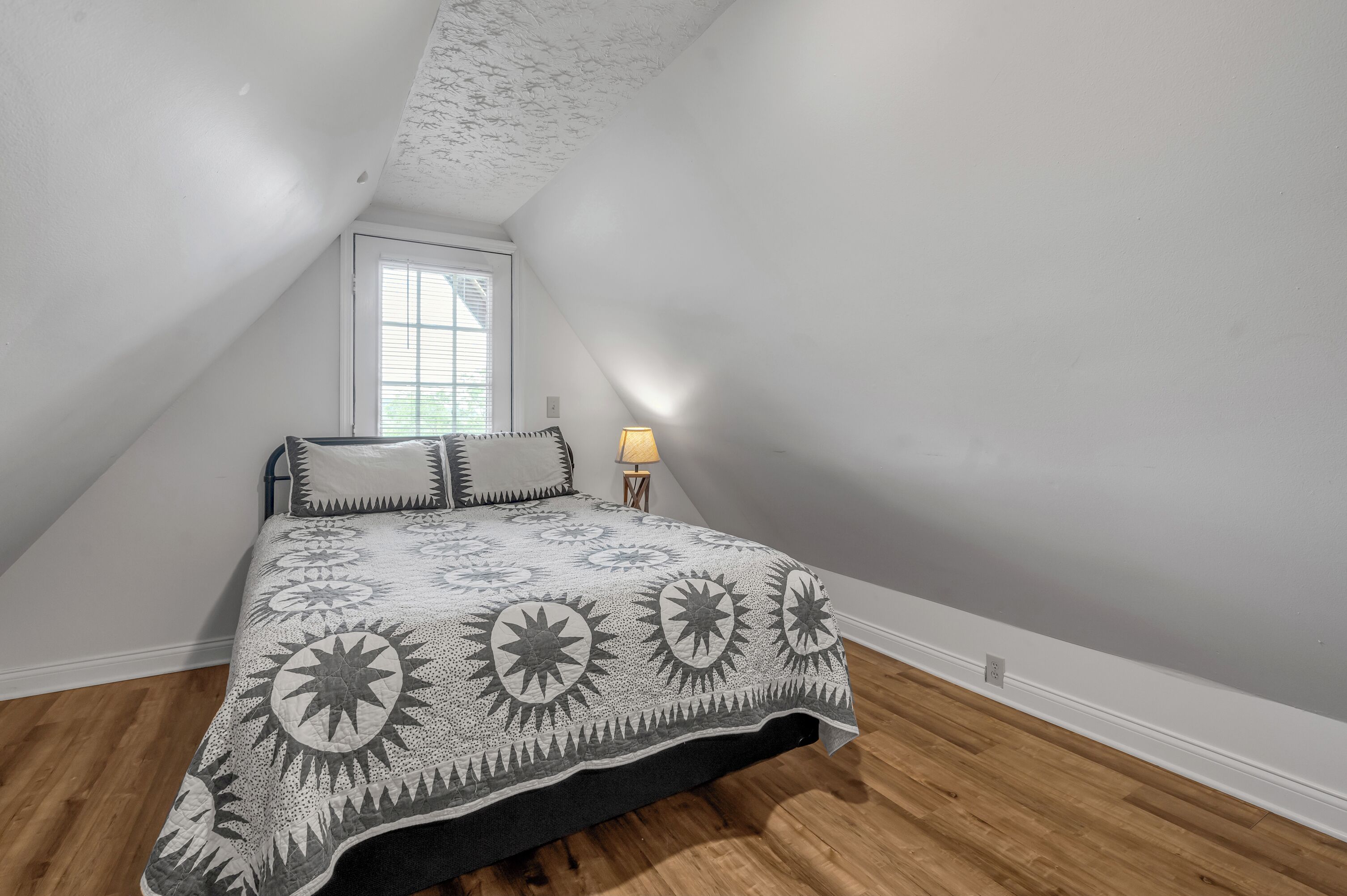 A bedroom with a sloped ceiling, a bed with a patterned bedspread, a bedside lamp, and a window with blinds.