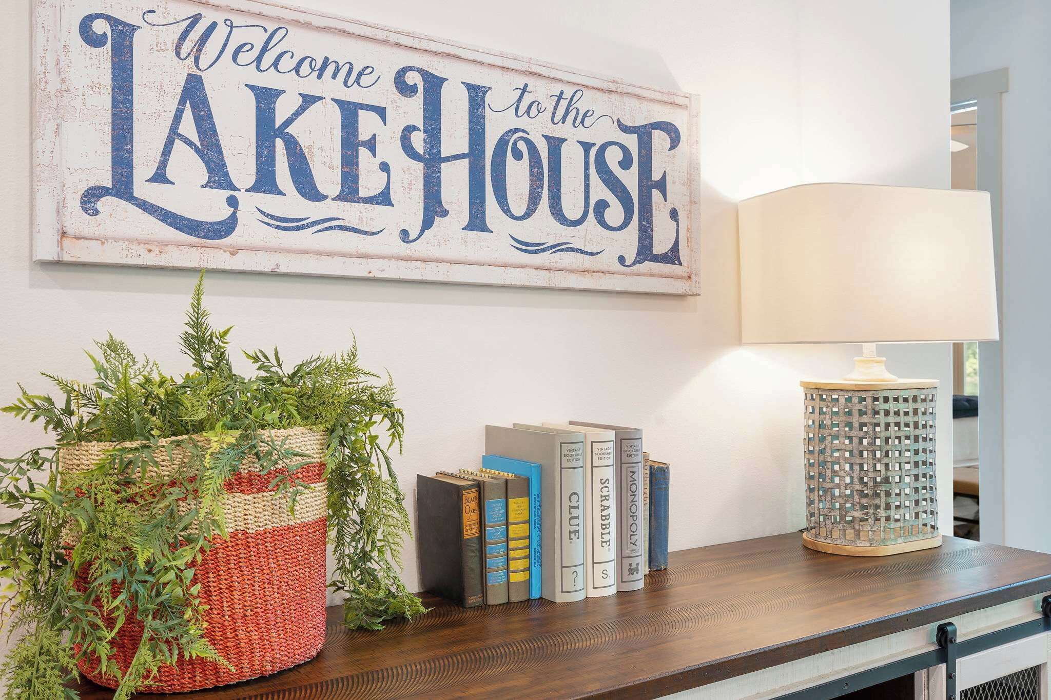 A welcome sign, a potted plant, a lamp, and books on a wooden table.