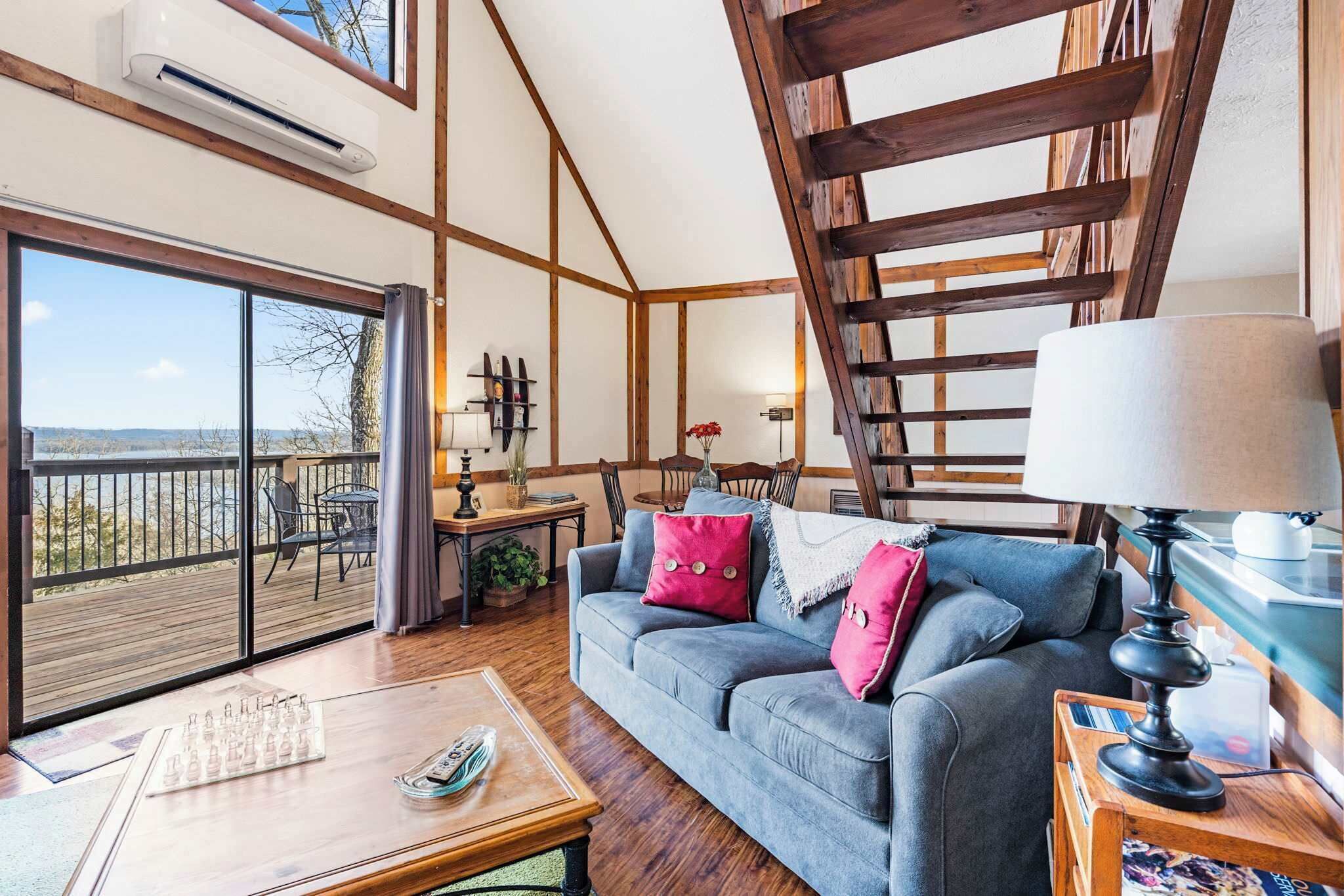 A cozy living room with a large window, a wooden staircase, and a balcony with a view.