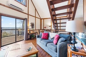 A cozy living room with a large window, a wooden staircase, and a balcony with a view.