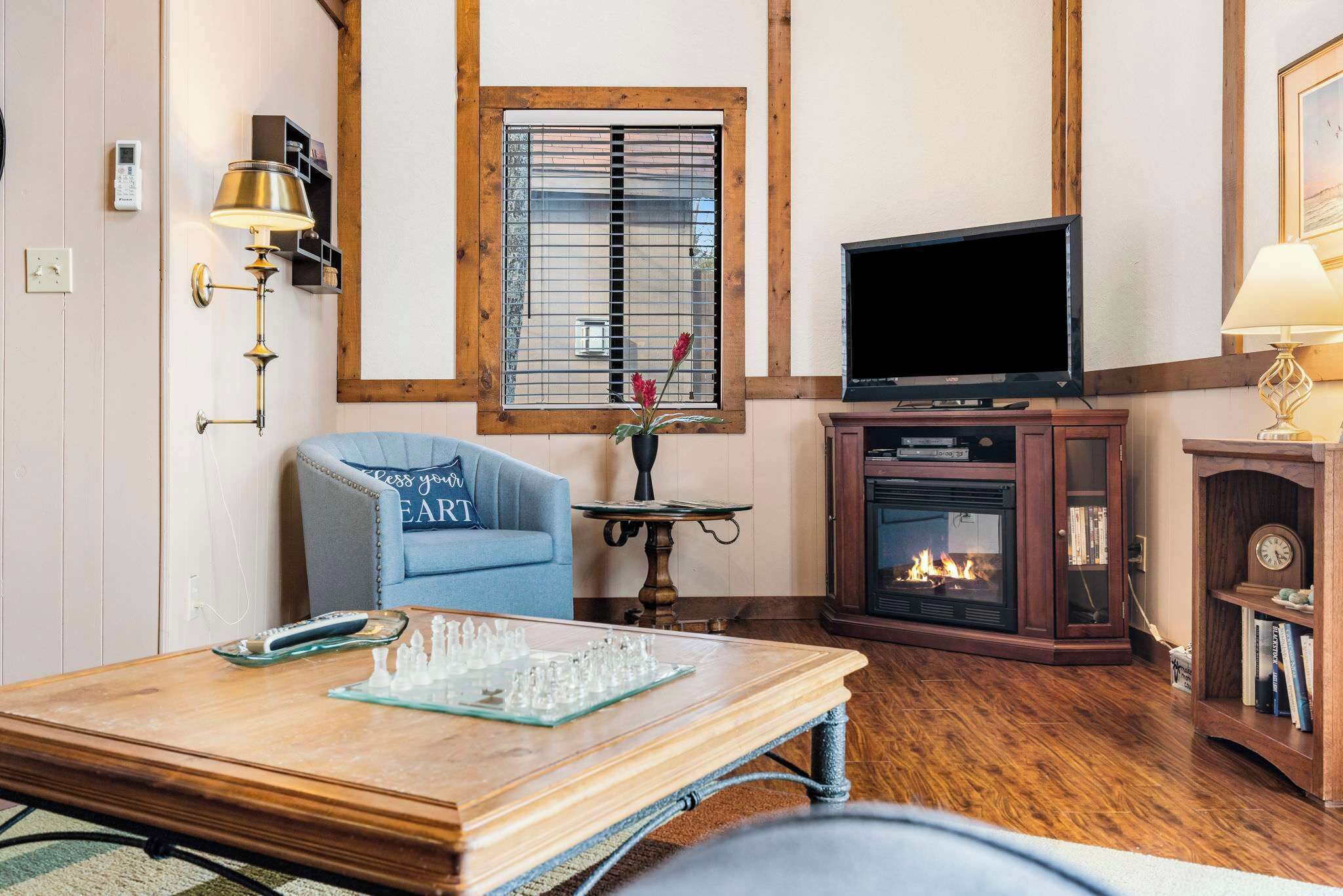 A cozy living room with a blue armchair, a wooden coffee table with a chess set, a fireplace, and a television.