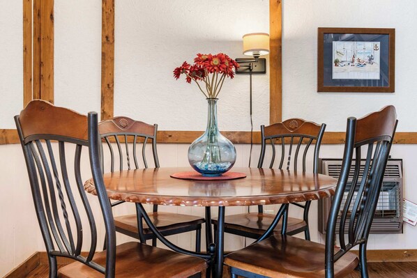 A wooden dining table with four chairs, a vase with red flowers, a wall-mounted lamp, and a framed picture on the wall.