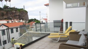 Een balkon met rieten meubilair, een glazen balustrade en uitzicht op gebouwen en een klif.