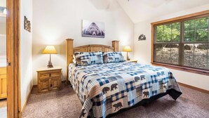 Un dormitorio con una cama grande, cabecera de madera, dos mesitas de noche con lámparas, ventilador de techo, ventana con persianas y un cuadro colgado en la pared.