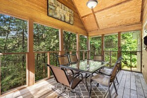 A covered outdoor deck with a glass table and chairs, surrounded by wooden beams and large windows offering a view of a forested area. - Baldknobber Ridge Escape | West Branson Cabin! (Branson West)