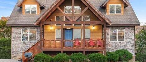 A two-story log cabin with a stone foundation, a wooden front porch with red chairs, and a stone chimney.