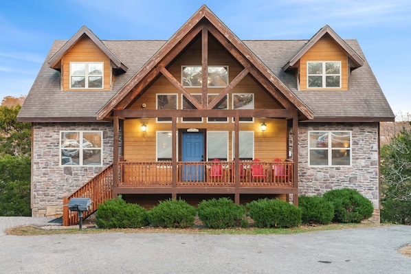 A two-story log cabin with a stone foundation, a wooden front porch with red chairs, and a stone chimney.