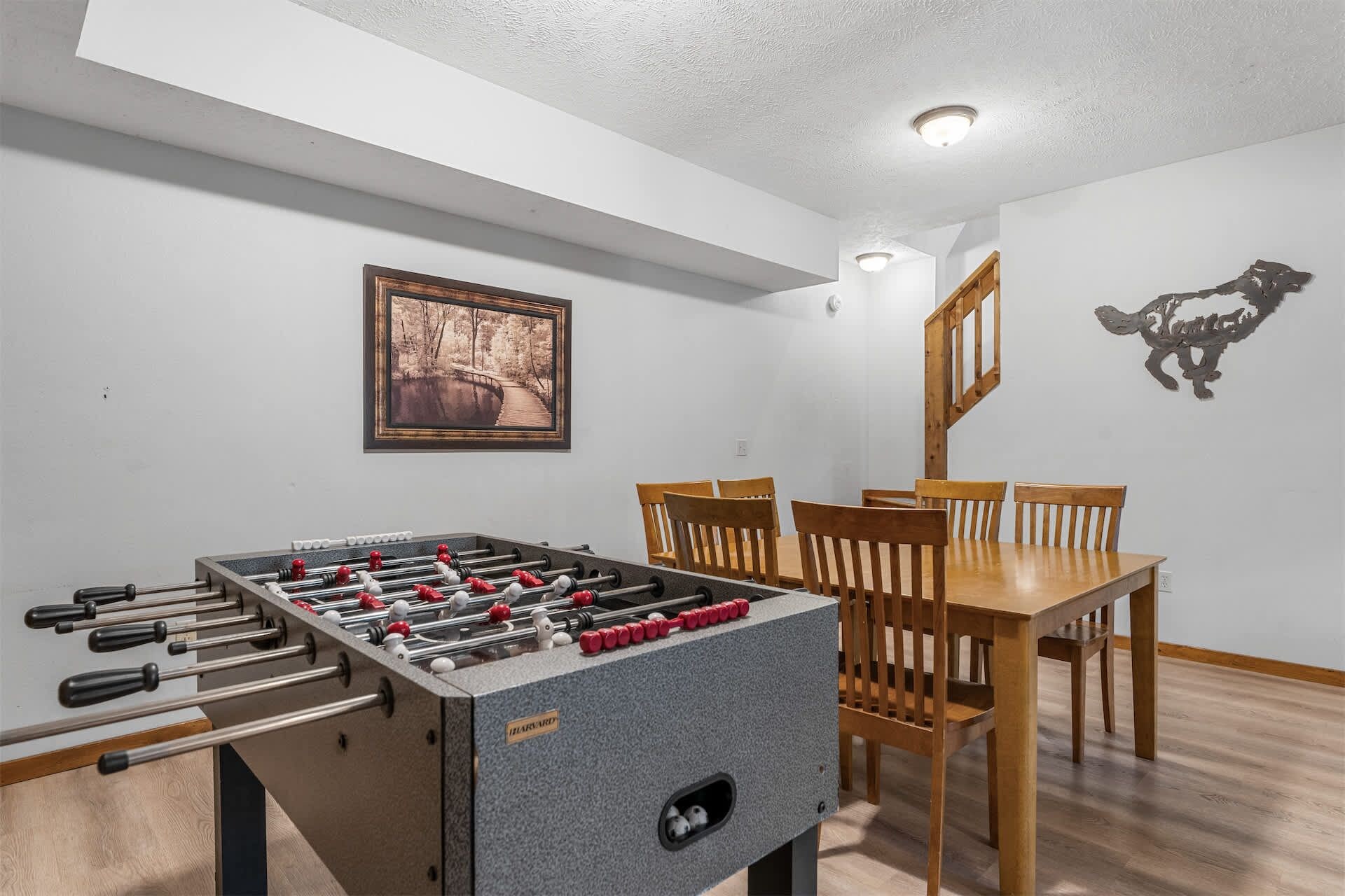 Een kamer met een tafelvoetbalspel, houten stoelen, een eettafel en een ingelijste foto aan de muur.