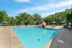 Une piscine avec un toboggan, des chaises longues, des parasols et des arbres en arrière-plan.