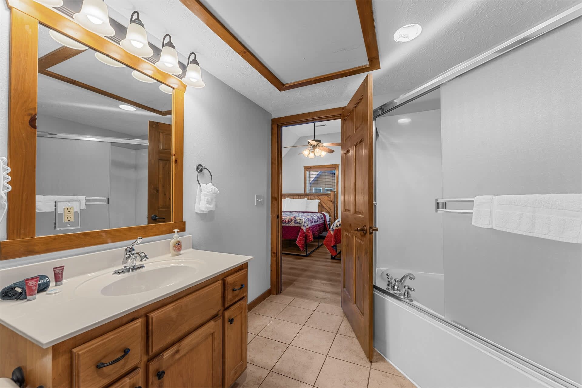 A bathroom with a large mirror, two sinks, a shower, and a door leading to a bedroom.