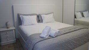 A neatly made bed with a grey blanket and pillows, a white bedside table with a vase, and a large mirror on the wall. - Villa Supranee relaxation a unforgettable stay, brand new  accommodation 850sft. (camara de lobos)