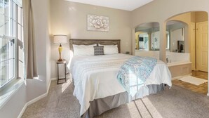 A bedroom with a large bed, a ceiling fan, a window with curtains, a bedside lamp, a wall-mounted flower artwork, a bathroom with a bathtub, and a mirror. - Holiday Ridge Escape | Holiday Hills Branson | Golf Resort | Outdoor pools (Branson)