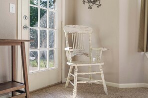 A room with a white wooden chair, a wooden table, and a window with a view of greenery outside.