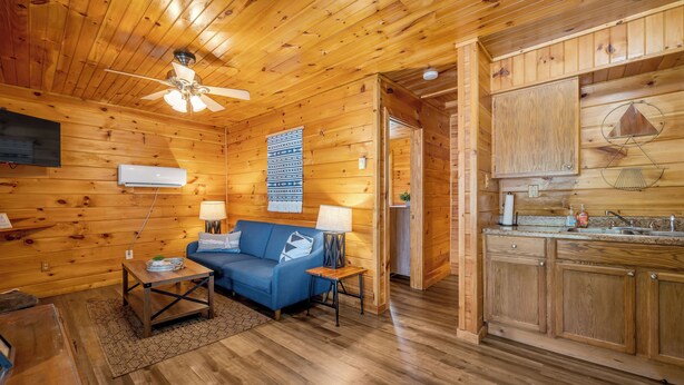 Un interior de madera con ventilador de techo, un sofá azul, una mesita y una zona de cocina.