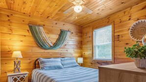 Un dormitorio de cabaña de madera con una cama, una mesita de noche, una lámpara, un ventilador de techo y una ventana con persianas.