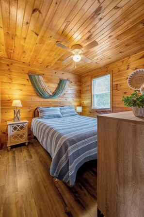 A wooden cabin bedroom with a bed, a nightstand, a lamp, a ceiling fan, and a window with blinds.