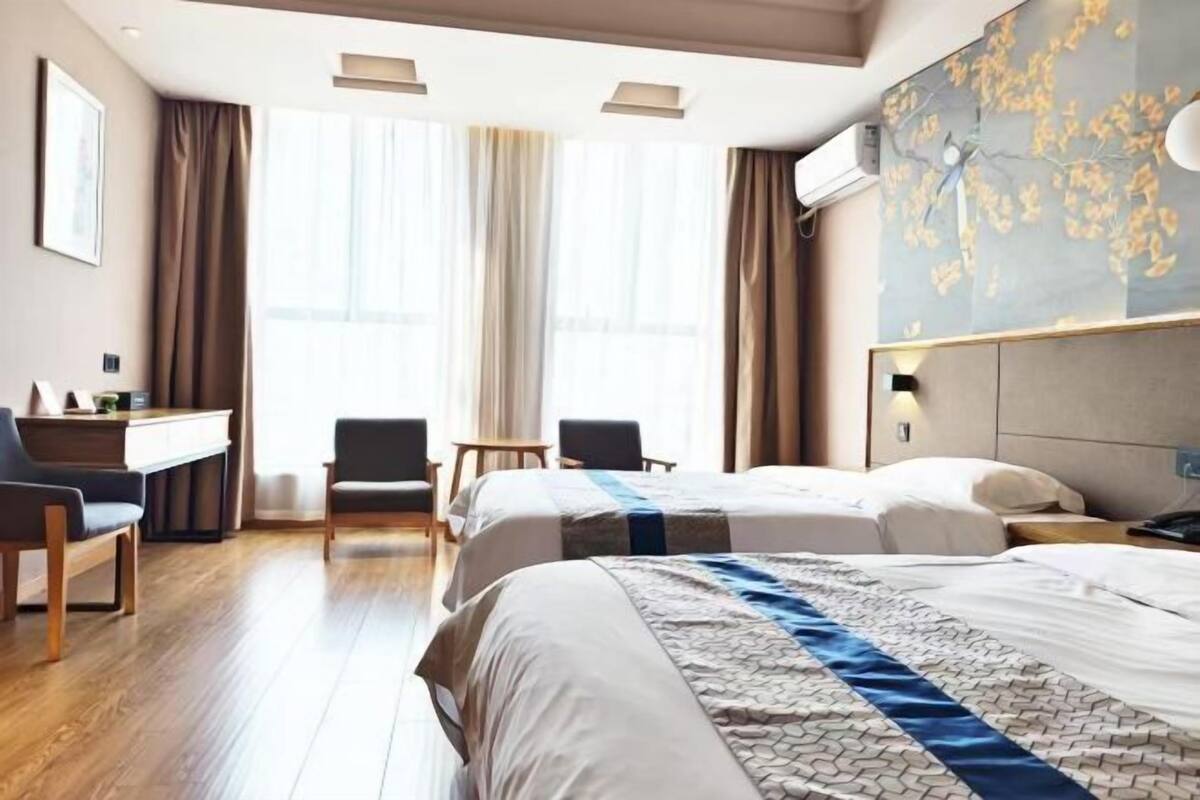 A hotel room with two beds, a desk, chairs, and a large window with curtains.