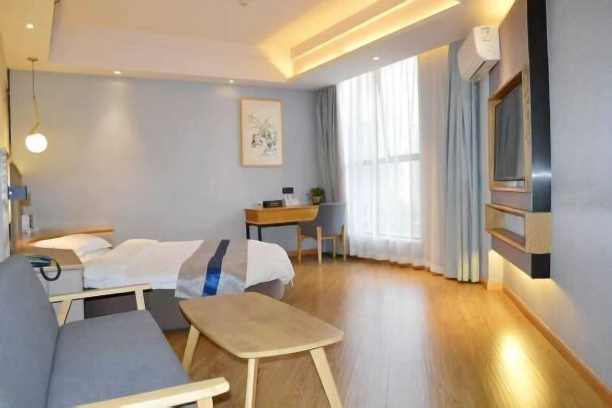 A modern hotel room with a bed, sofa, desk, and a large window with curtains.