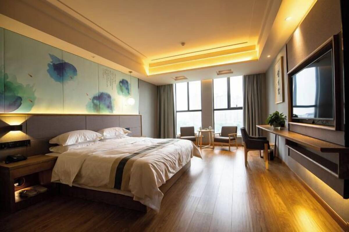 A modern hotel room with a large bed, bedside tables, a flat-screen TV, a seating area with chairs, and a large window.