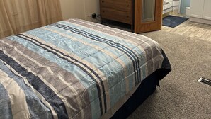 Un dormitorio con una cama, un aparador con espejo, un ventilador y una puerta que da a otra habitación.