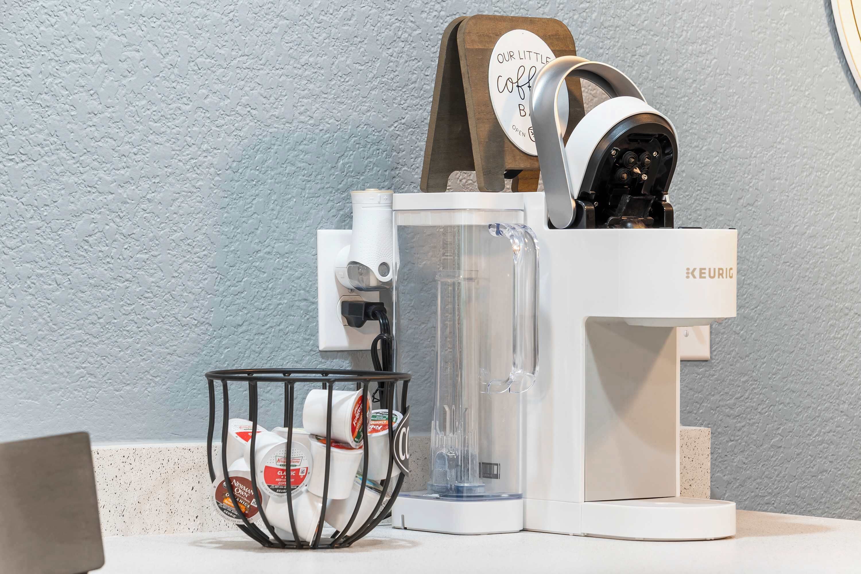 A coffee machine with a clear water tank, a coffee pod holder, and a sign indicating 'Our Little Coffee Bar' is open.