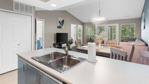 A modern kitchen with a double sink, a dining area with a table and chairs, and a living room with a sofa and a TV. - Linda’s Haven – Golf Course View (Branson)