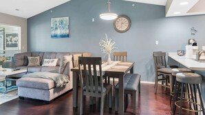 A modern living room with a gray sofa, a dining table with chairs, a kitchen area, and a large clock on the wall. - Linda’s Haven – Golf Course View (Branson)