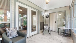 A covered patio area with a ceiling fan, a seating area with a cushioned chair and a table, and a decorative wall clock. - Linda’s Haven – Golf Course View (Branson)