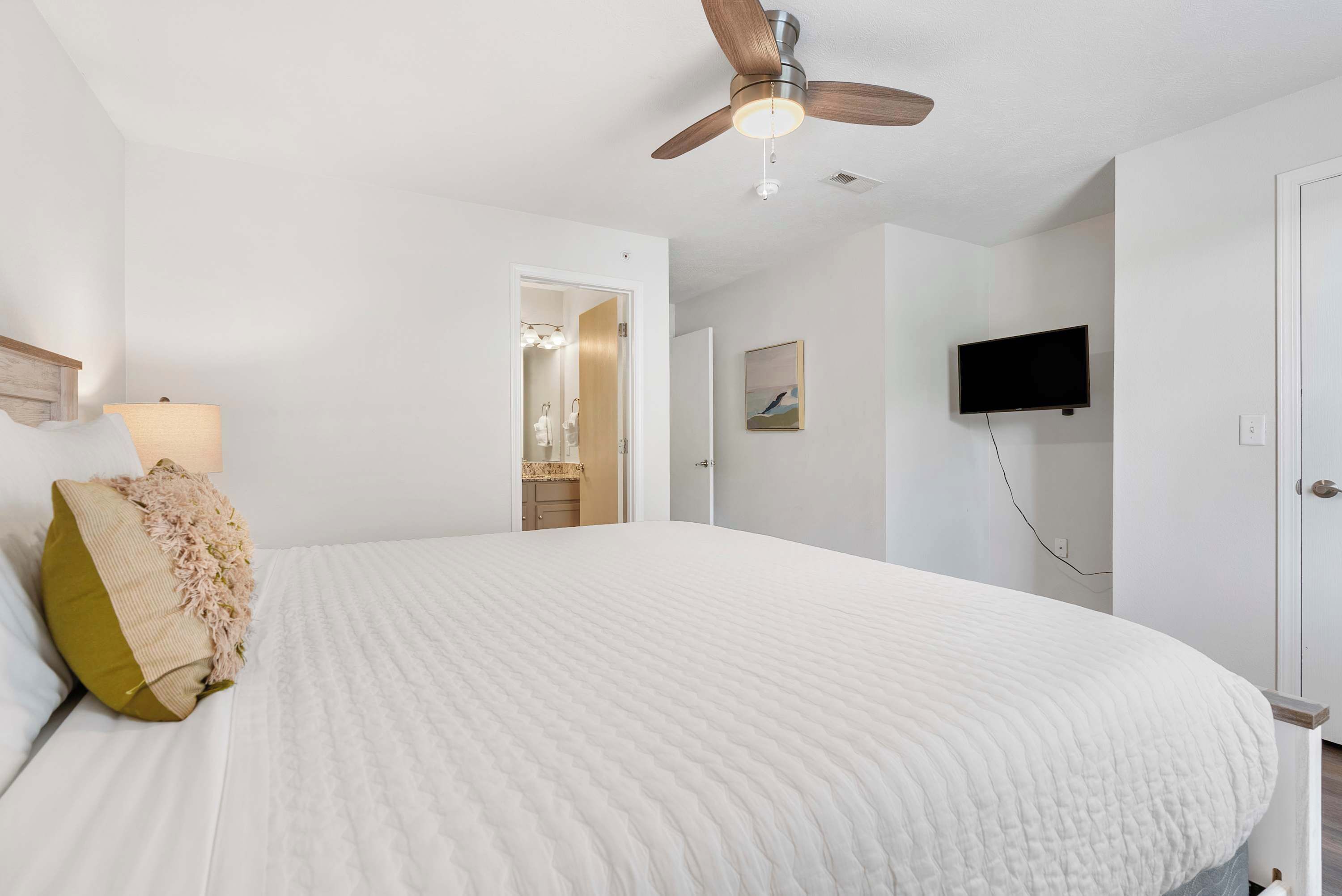 A bedroom with a large bed, a ceiling fan, a TV mounted on the wall, and a door leading to another room.