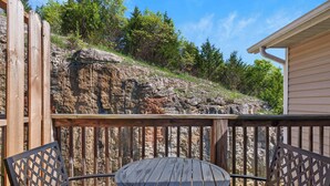 A wooden deck with a round table and two chairs, overlooking a rocky hillside and trees. - Hillside Cottage in The Vineyards Resort! (Branson)
