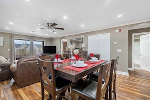 Un coin repas avec une table en bois dressée pour quatre personnes, un ventilateur de plafond et une vue sur l’extérieur grâce à de grandes fenêtres.