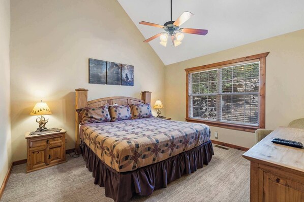 Une chambre à coucher avec un lit, deux tables de chevet, un ventilateur de plafond, une fenêtre avec des stores et un tableau au mur.