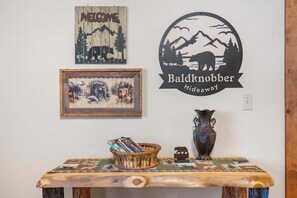 Een houten tafel met een mand tijdschriften, een lijst met een afbeelding, een decoratieve vaas en een bord met een bergsilhouet.
