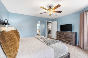 A bedroom with a bed, a ceiling fan, a dresser, a TV, and a door leading to another room.