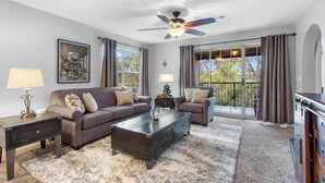 A living room with a dark wood coffee table, two sofas, a ceiling fan, and large windows with curtains.