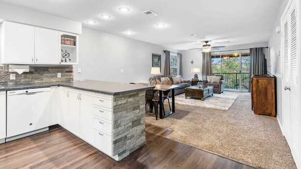 A modern kitchen with white cabinets, a stone island, and recessed lighting. There is a dining area with a table and chairs, a sofa, and a view of the outdoors.