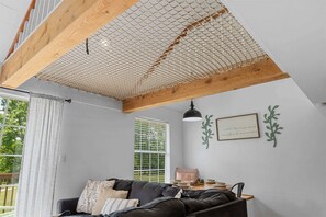 A room with a black leather sofa, a dining table with dishes, a window with blinds, and a ceiling with a wooden beam and mesh.