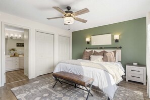 Quarto com cama, um banquinho, mesa de cabeceira, ventilador de teto e espelho.