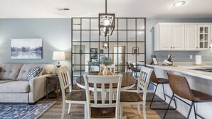 Una sala de estar moderna con sofá, mesa de comedor y sillas. Cuenta con una zona de cocina con armarios y encimera.