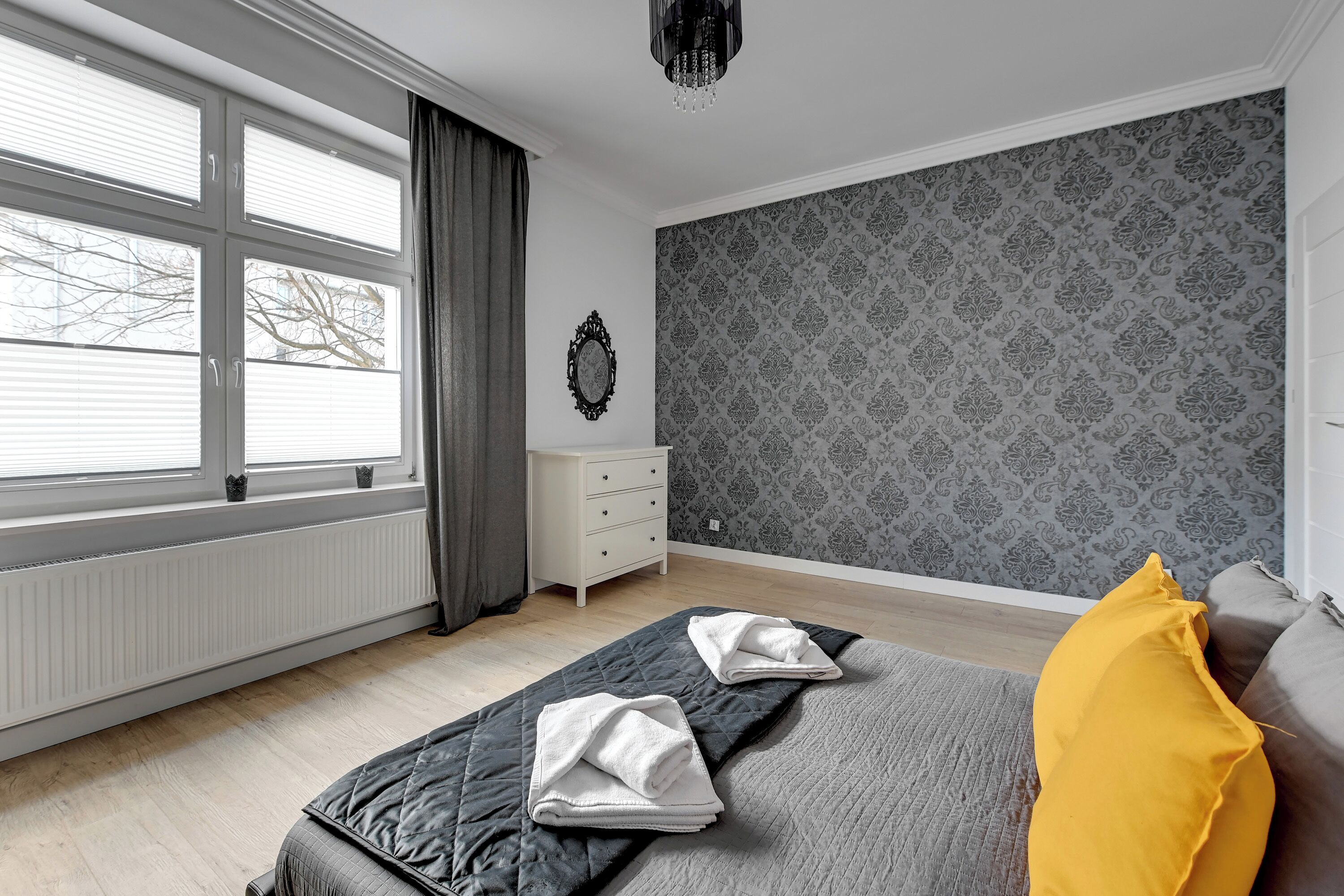 A bedroom with a bed, a nightstand, a dresser, a window with curtains, and a patterned wallpaper.