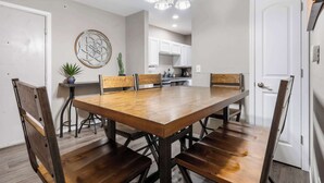 A dining area with a wooden table and chairs, a chandelier, and a kitchen in the background.