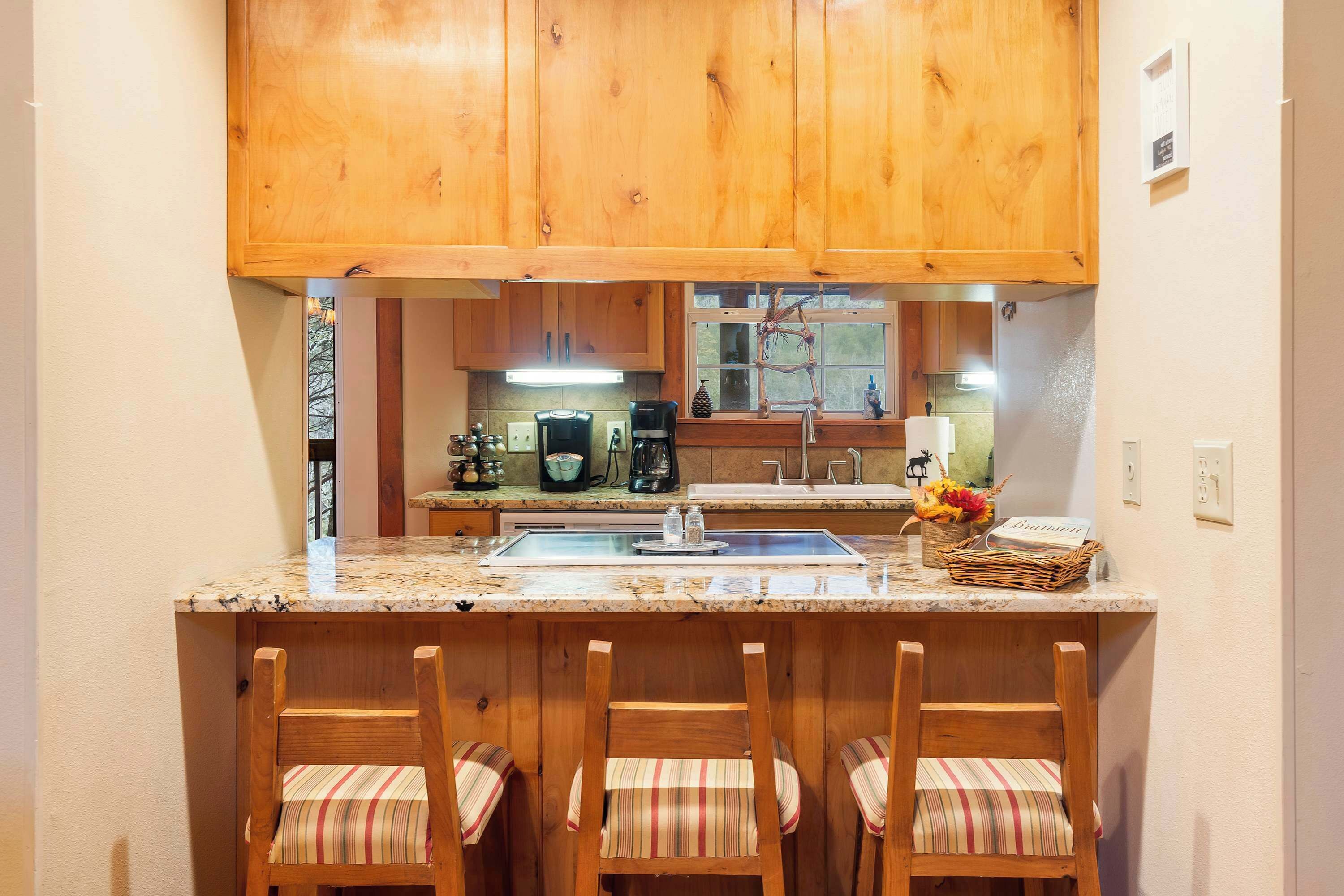 A kitchen with wooden cabinets, a granite countertop, a coffee maker, and a sink. There are three striped chairs at the counter.
