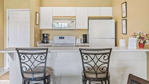 Une cuisine avec des armoires blanches, un four à micro-ondes, une cuisinière et un réfrigérateur. On y trouve deux chaises avec des structures en métal et des assises rembourrées.