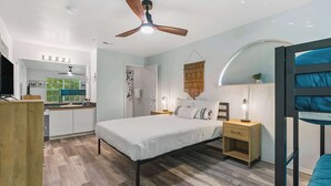 Une chambre à coucher avec un lit, des tables de chevet, un ventilateur de plafond et une vue sur la cuisine.