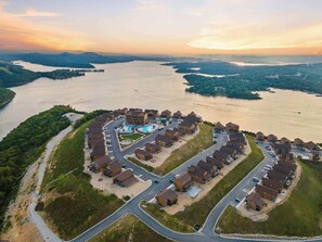 A resort with a central pool, surrounded by single-story houses with parking, set against a backdrop of a large lake and distant hills. - A Birds Eye Bridge View – 5 BDR w/ Private Hot Tub (Kimberling City)