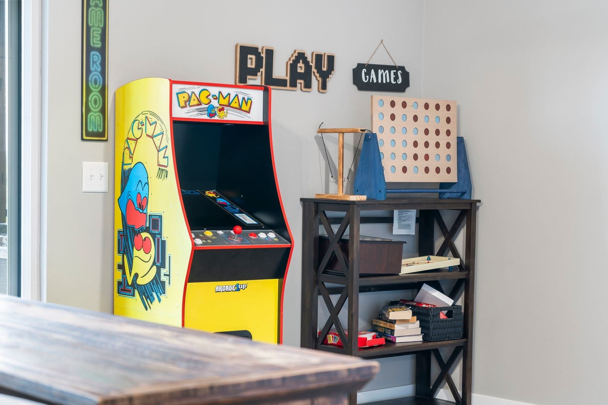 Ein Zimmer mit einem Pac-Man-Spielautomaten, einer Holzwand mit Spielsteinen und einem Bücherregal mit Büchern und Spielen.