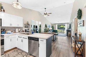 A modern kitchen with white cabinets, a dishwasher, and a large island. There is a ceiling fan, a dining area with a table and chairs, and a view of the outdoors. - Vansen's Golfshores – Inviting, Cozy Condo on Golf Course! (Branson)