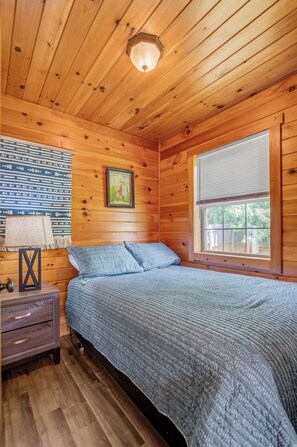 Dormitorio de cabaña de madera con una cama, una mesita de noche, una lámpara, un cuadro enmarcado y una ventana.
