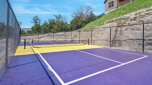 A tennis court with a net, surrounded by a chain-link fence, and a building with a wooden deck in the background. - Two Pines – Endless Views and Attractions! (Branson)