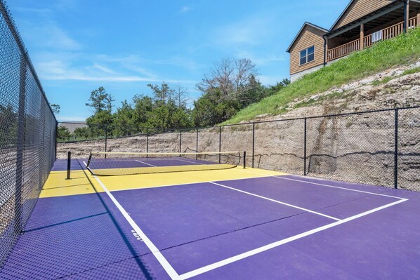 A tennis court with a net, surrounded by a chain-link fence, and a building with a wooden deck in the background.